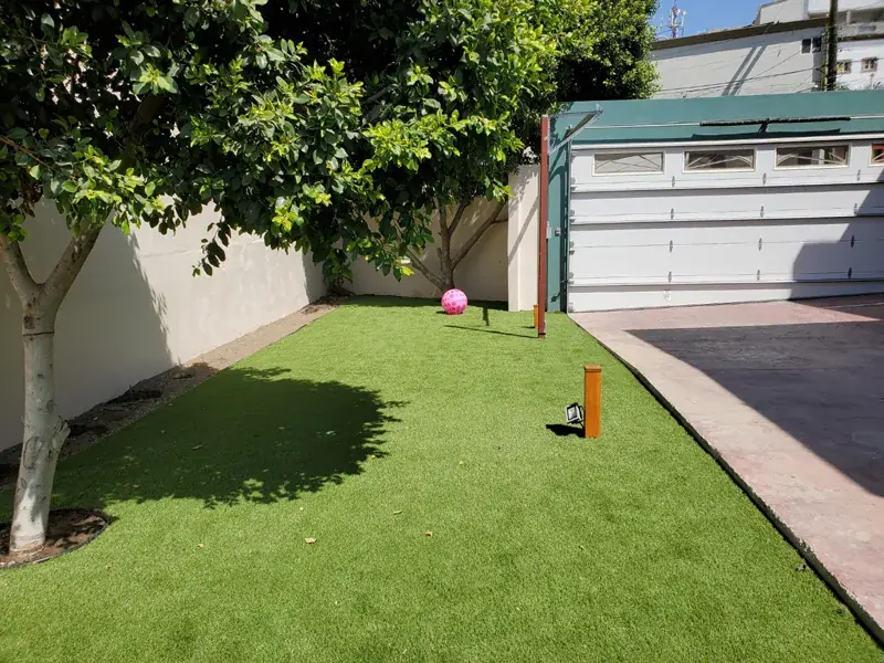 Jardín con árbol de sombra sobre pasto sintético, junto a la cochera