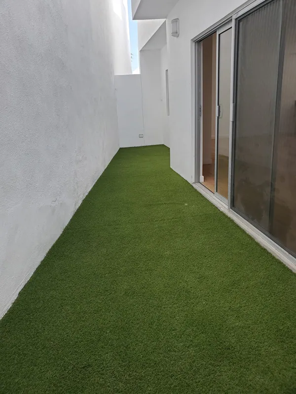 Pasillo lateral angosto entre muros blancos cubierto con pasto sintético